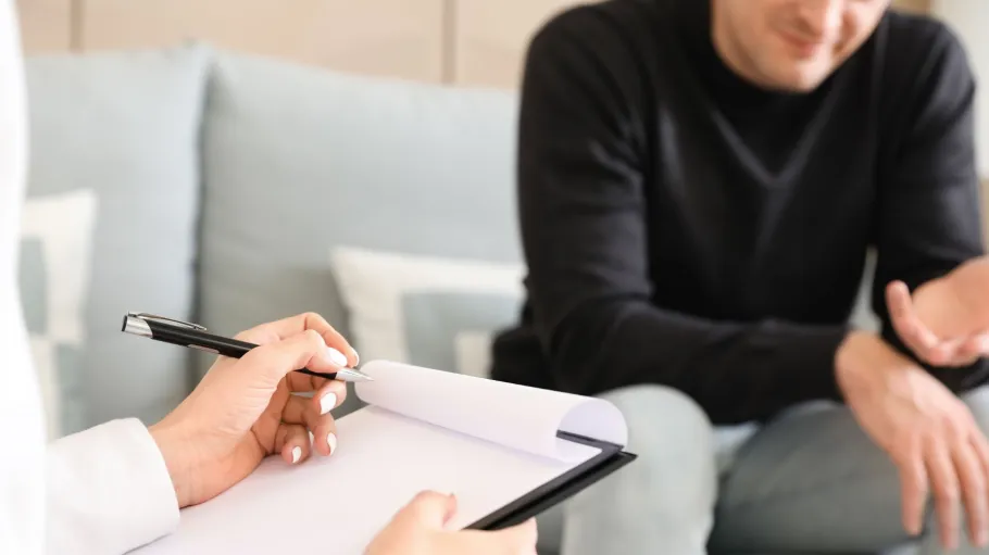 A close up of a counselling appointment. In the foreground, someone is taking notes. In the background, someone is sitting on a couch, gesturing with their hands while talking.