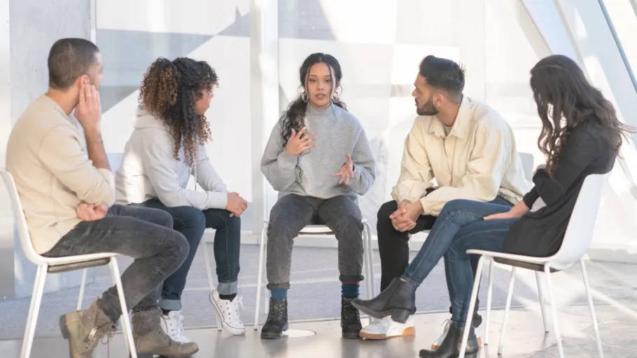 Five students sit on chairs in a half circle formation. Four look towards the woman in the middle, who is speaking and gesturing with her hands.