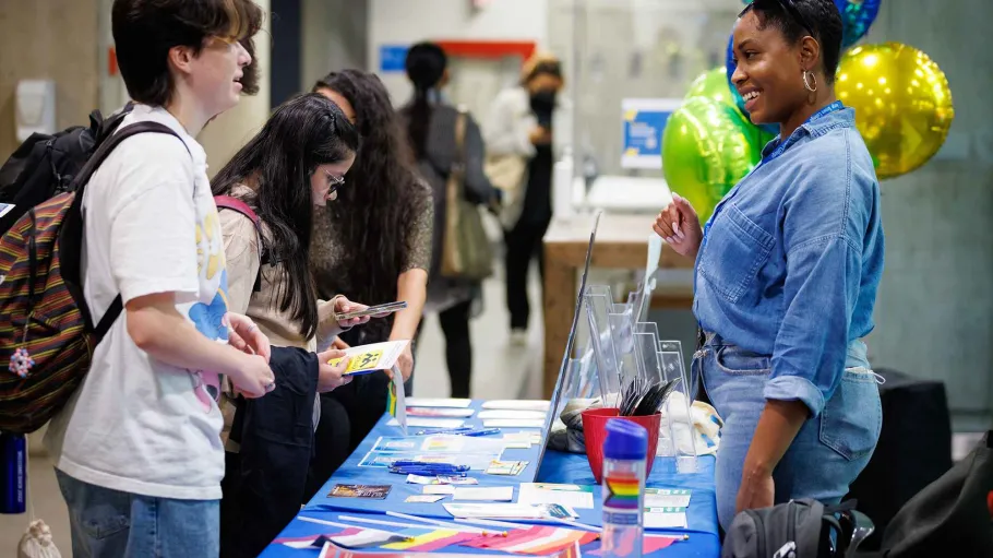 A student stands in front of a table at a fair, talking to the volunteer behind the table. Other students in the background examine what's on the table.