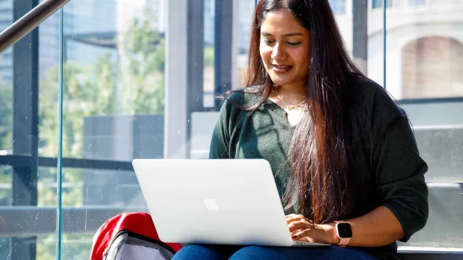 A student sits on stairs in front of a window as she works on her laptop.