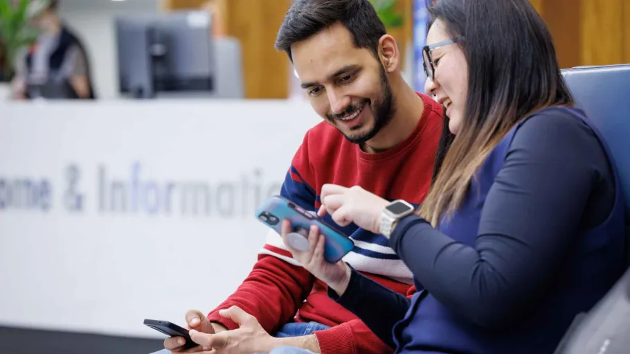 Two students sit in front of an information booth. One points at their phone while the other looks over their shoulder at it.