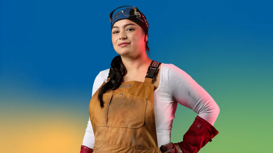 A woman in coveralls, safety glasses, and welding gloves smiles and looks confidently ahead against a colourful blue background.