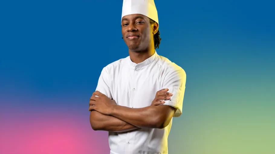 A Black man in chef whites crosses his arms and looks directly ahead, while standing in front of a colourful blue gradient background.