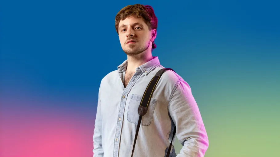A man with short hair and beard wearing a denim shirt looks confidently ahead.