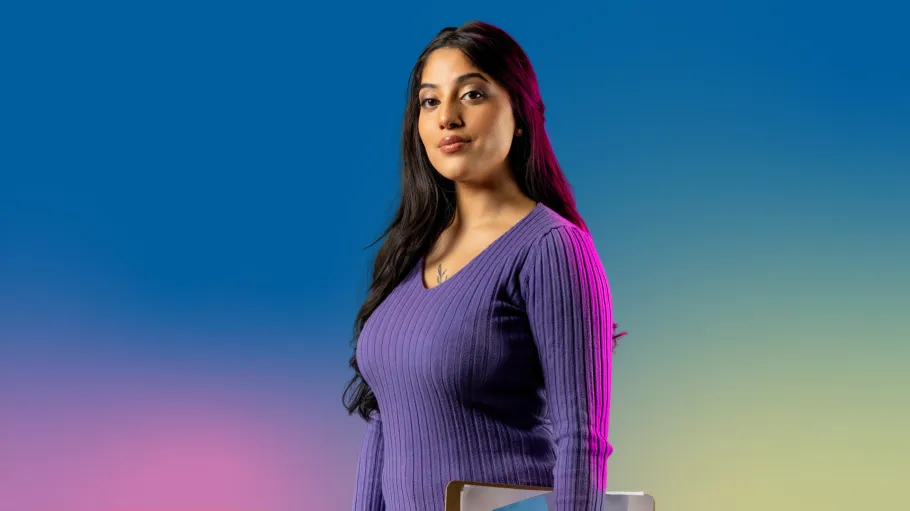 A woman in a purple sweater with long dark hair holds a clipboard under one arm and looks confidently ahead.
