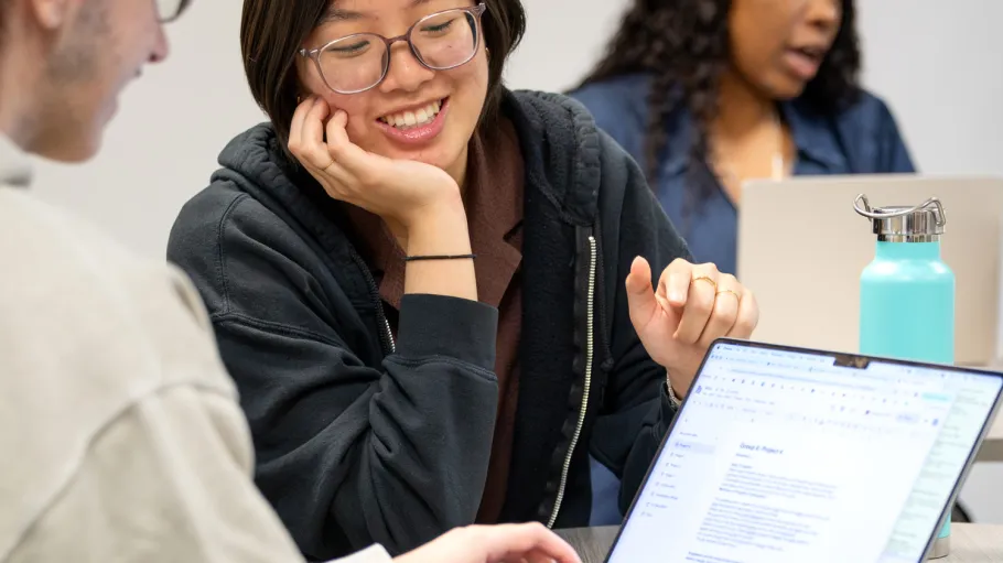 Two students seated at a table looking at a laptop screen while one types away.