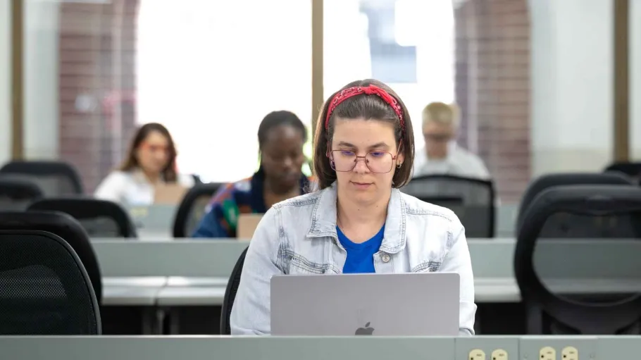 A student works at her laptop. Out of focus behind her are more students doing the same.