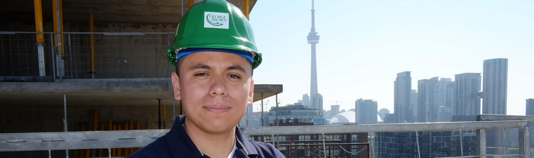 A male construction management student poses at a high ground worksite with the backdrop of the Toronto skyline behind him. Student is smiling and has arms crossed to show confidence.