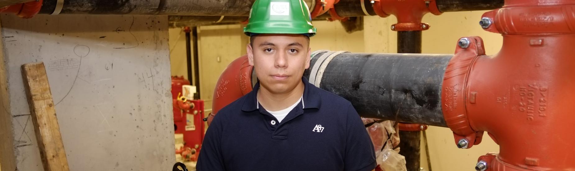 A male construction management student poses in a room with large red pipes while holding an architectural diagram and wearing a green hardhat.