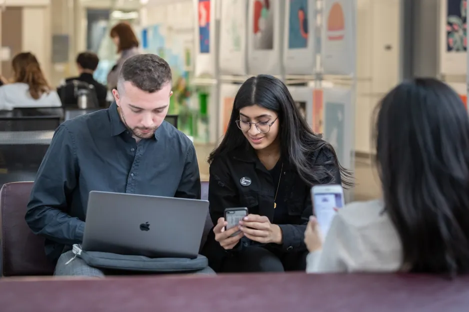 Two students sitting beside each other, one looking at a computer, the other at a phone.