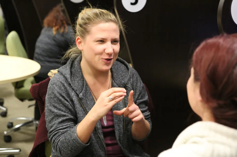 Female American Sign Language and Deaf Studies student practices American Sign Language with another female student seated at video monitor in ASL lab cubicles.