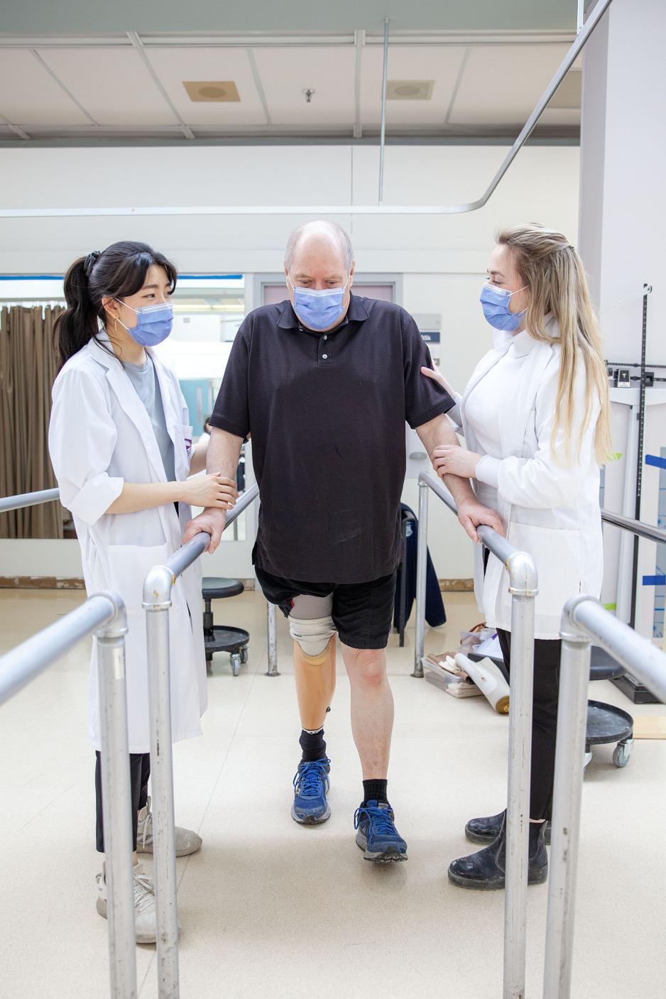 Two female Clinical Method in Orthotics/Prosthetic (S407) students work with an elderly male client as he learns to walk with his new prosthetic limb.