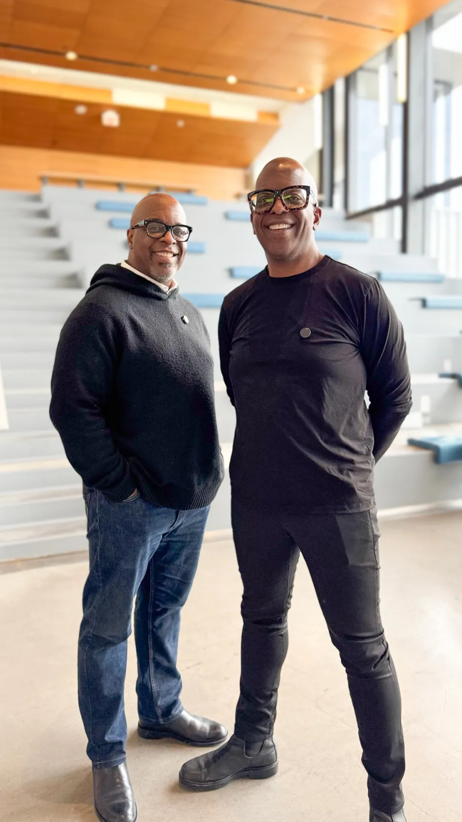 Two men in dark shirts and dark framed glasses smile