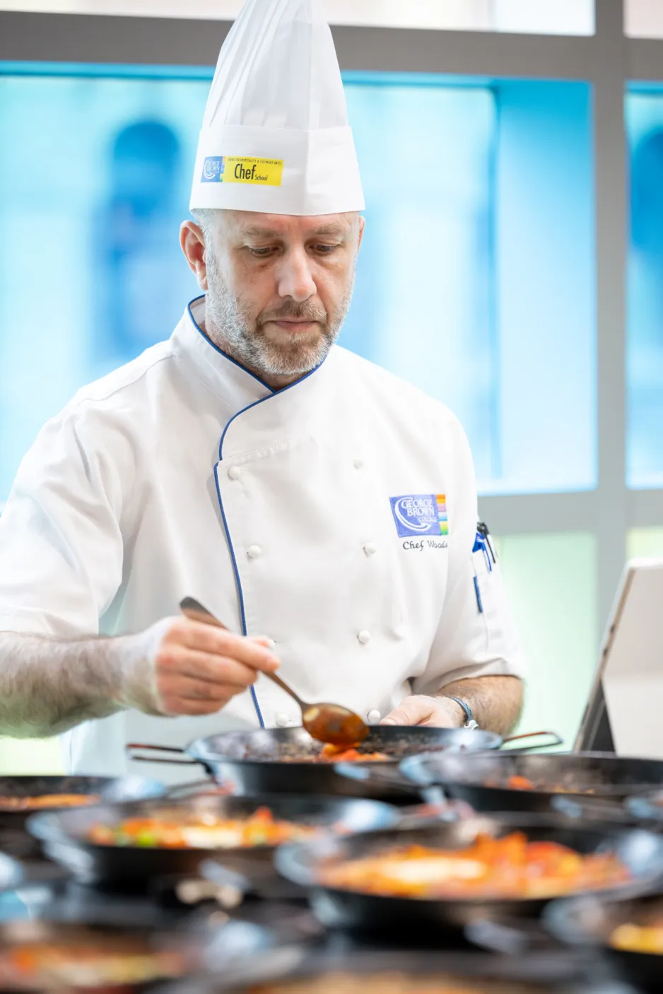 A man in chef whites works over stirs a dish in a busy kitchen