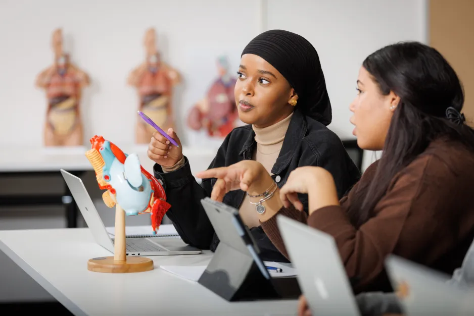 Two students chat at a desk with their laptops open