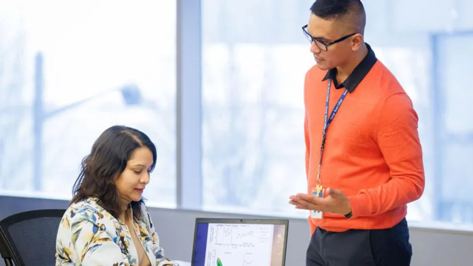 Man and woman studying Behavioural Science Technology Advanced Diploma program. A woman sits at a table with a laptop while a standing man speaks to her.