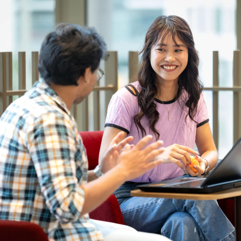 Two seated students chat in front of an open laptop