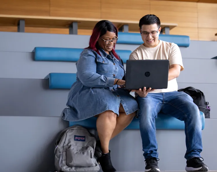 Two students are seated in a learning landscape looking at a laptop.