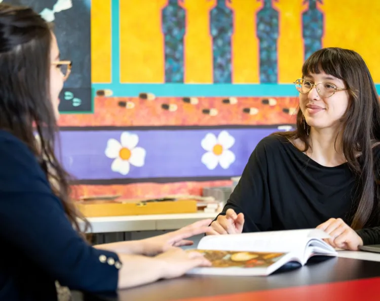 Two students in the Indigenous Initiatives office sit at a table in discussion, with a book and laptop on the table. 