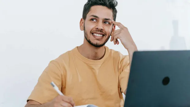 A student sits in front of a laptop, also holding a pen to a notebook as if writing. He's looking to the side with his finger on his temple as if he's in thought.