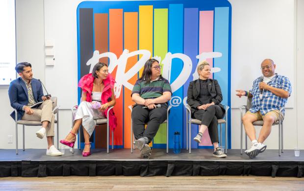 A group of panelists sits on a stage in front of a large colourful background with the word 'Pride'