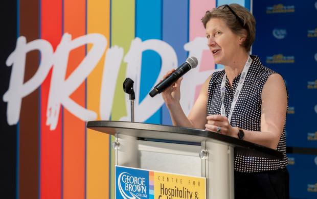 Karine Morin speaks into a microphone at a podium with a colourful pride banner in the background