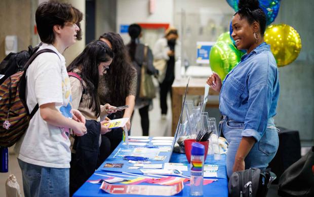 A student stands in front of a table at a fair, talking to the volunteer behind the table. Other students in the background examine what's on the table.
