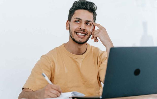 A student sits in front of a laptop, also holding a pen to a notebook as if writing. He's looking to the side with his finger on his temple as if he's in thought.