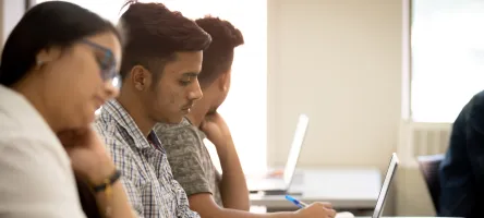 Two male students and one female student taking notes in class with a laptop and paper and pen.