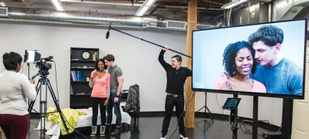 Female and male acting student are in front of the camera in a video studio and being recorded while acting. Closeup of their faces is visible on large TV screen.