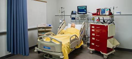 imulation Centre. The room has a dummy laying on a hospital bed, in a hospital room. There is a cabinet on the right hand side of the photo. Above the hospital bed is a monitor.