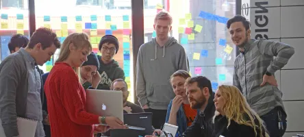 Group of people sitting and standing around a table during a meeting while someone is presenting.