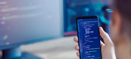 A hand holding a mobile phone with computer code on it in the foreground with a desktop computer in the background.