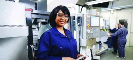Smiling female mechanical Engineering student poses in front of machine holding tool.