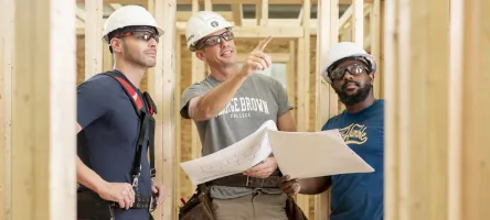 Three people in safety equipment and hard hats look at construction drawings and wood framing