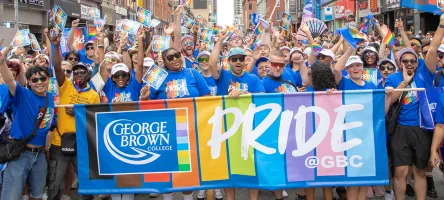 A large group of people in blue shirts cheer while holding a Pride @ GBP banner