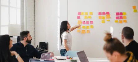 Group of people in a meeting room, with one person leading a discussion and pointing to sticky notes on a wall.