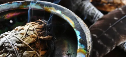 A close up image of a burning white sage smudge stick, healing crystals, and sacred feather. 