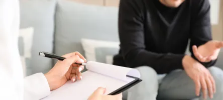 A close up of a counselling appointment. In the foreground, someone is taking notes. In the background, someone is sitting on a couch, gesturing with their hands while talking.