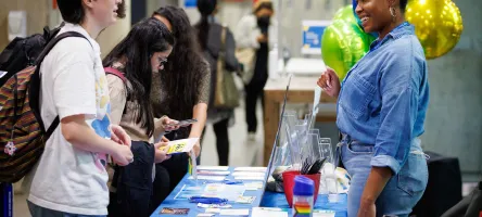 A student stands in front of a table at a fair, talking to the volunteer behind the table. Other students in the background examine what's on the table.
