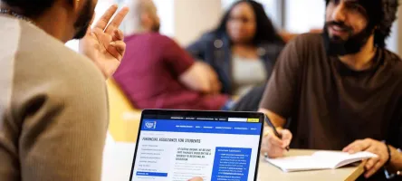 Two students sit across from each other. One's laptop is facing the camera, open to the Financial Assistance page.